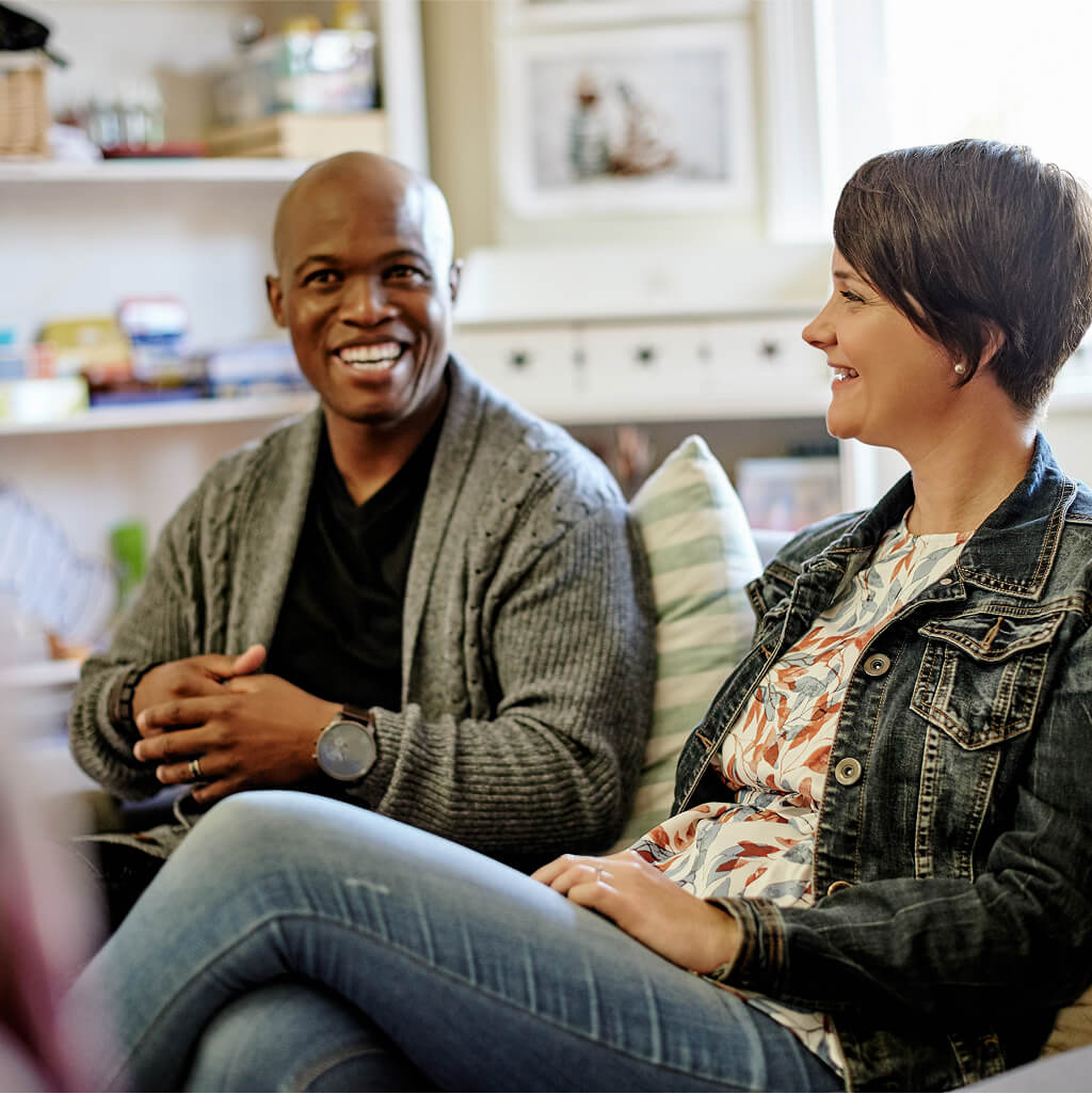 Two people sit indoors on a couch, smiling and having a conversation about art therapy. Shelves and framed pictures are visible in the background.