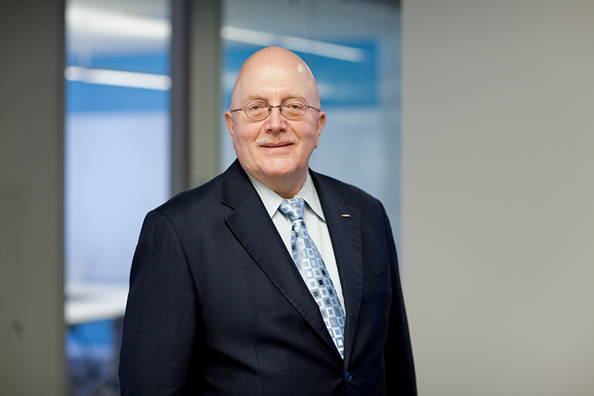 A bald man wearing glasses, a dark suit, a light blue shirt, and a patterned tie stands in an office setting, smiling at the camera—Joseph Troiani, author of A City at War: Chicago WWII.