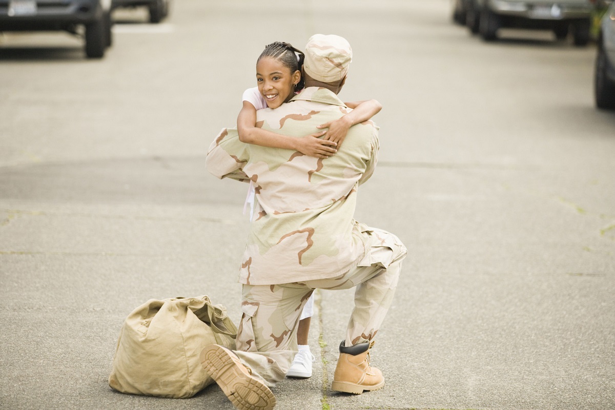 A person in military uniform kneels and embraces a smiling child on a street, with a duffel bag placed nearby—a heartfelt moment often explored in military psychology research.