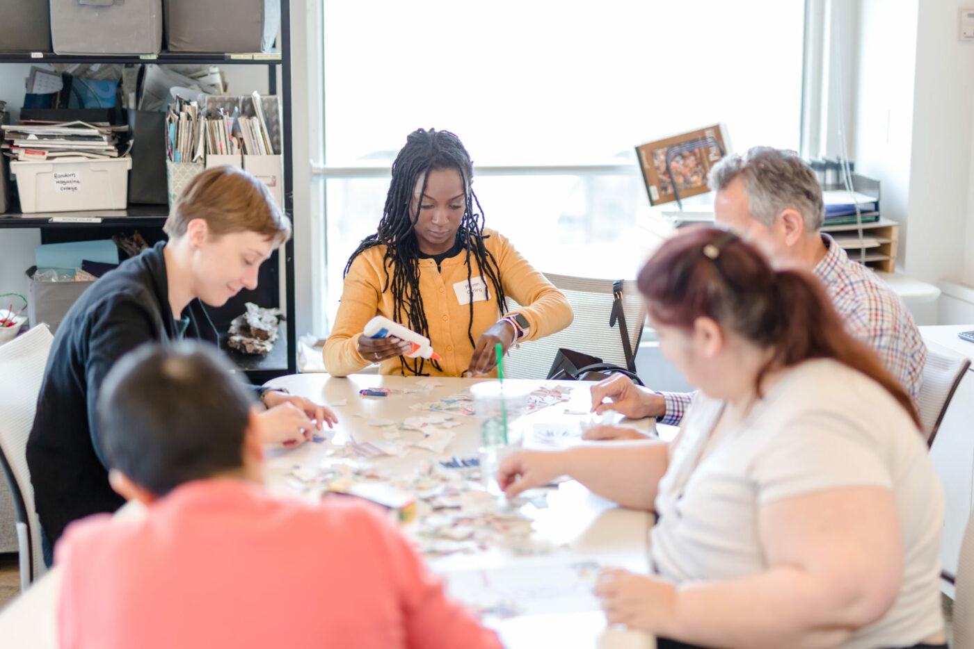 A group of five people sit around a table, working on an arts and crafts project with paper and scissors in a bright, organized room that reflects contemporary practice inspired by Adler’s principles.