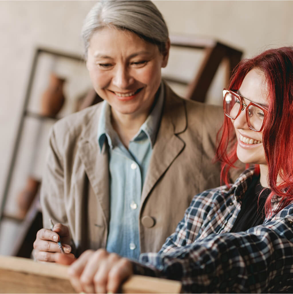 Two women stand together indoors, one with Grey hair and a beige jacket, the other with red hair and glasses—both smiling as they discuss M.A. studies in Art Therapy or Counselling Psychology.