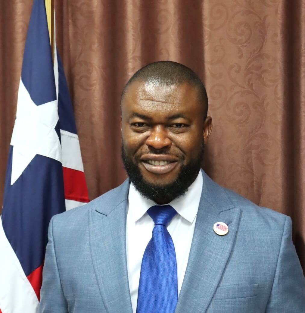 A man in a blue suit and tie stands in front of a brown curtain and a flag with a white star and stripes, smiling while wearing a round pin on his lapel—embodying transformational change within Liberia’s Civil Service.