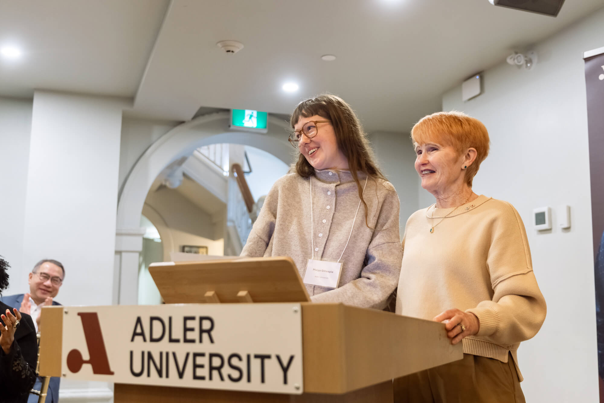 Two women stand together at a podium labeled "Adler University," smiling, with one holding a laptop. The scene reflects community care, with people visible in the background.