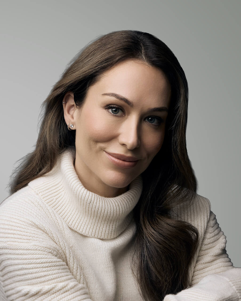 Carolyn Charest, a woman with long brown hair wearing a white turtleneck sweater, smiles softly while sitting against a plain grey background.