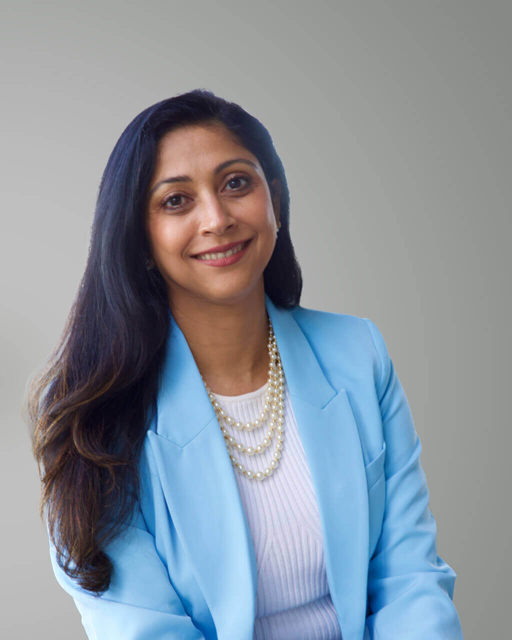 Woman with long dark hair wearing a light blue blazer, white top, and layered pearl necklace, smiling confidently against a plain light Grey background—balancing style and poise in today’s fast-paced AI workplace.
